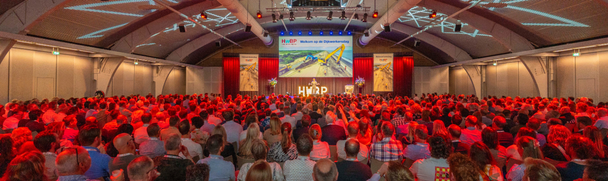 Een grote groep dijkwerkers in een zaal waar een spreker op het podium staat tijdens de Dijkwerkersdag 2025