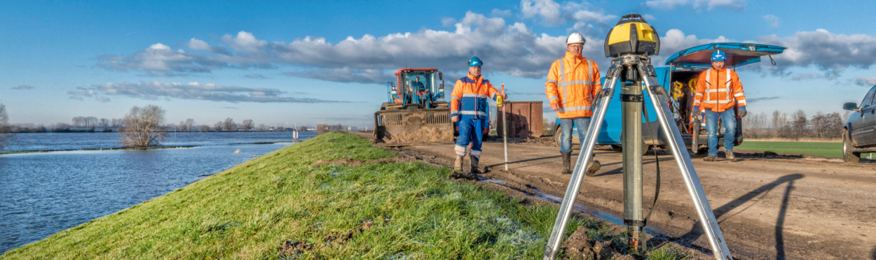 Drie dijkwerkers lopen je tegemoet. Op de voorgrond staat een meetinstrument.