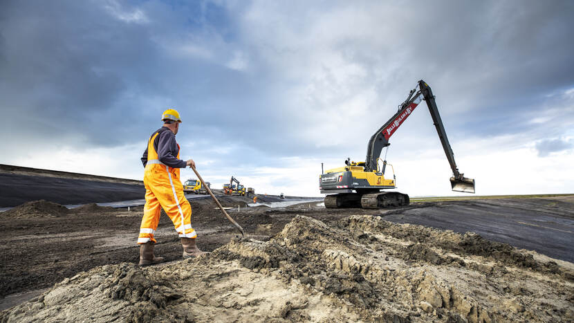 Man in overall staat met schep in de modder tijdens de uitvoering versterking Lauwersmeerdijk